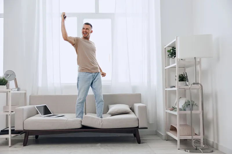 A joyful man enjoying a digital detox, dancing on the couch with a phone in hand, surrounded by a light and airy home environment, promoting mental well-being.