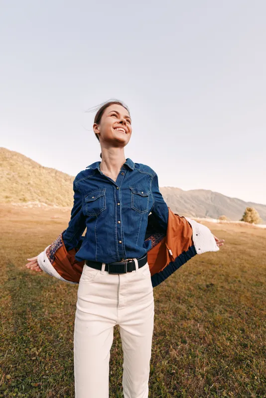 Woman with smile outdoors in a field wearing denim shirt and jacket, happy and carefree among mountains and grass, portrait of joy, sunlight and open arms in scenic landscape.