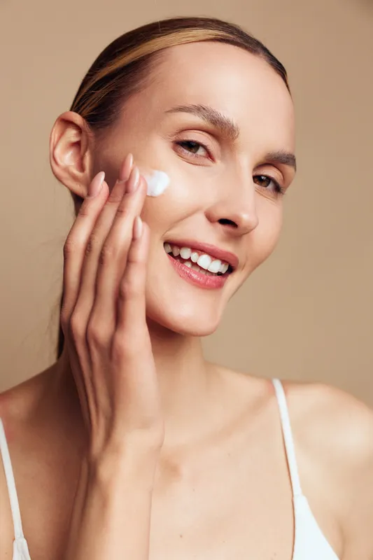 Smiling woman applies moisturizing cream on face with fingers showing skincare routine and healthy skin care concept isolated on beige background in closeup portrait.