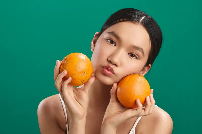 asian woman holds oranges in a studio setting with a vibrant background, offering a fresh, playful mood as she leans toward the camera.