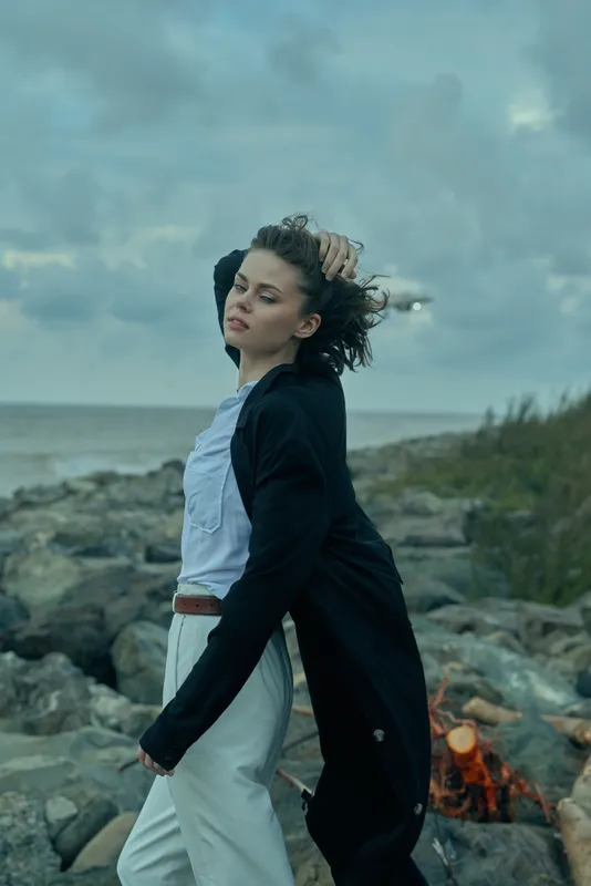 Woman standing on rocky coast, wind in hair, wearing a long black coat over a light shirt and white trousers, near a small campfire, expressing freedom and moody coastal fashion in an outdoor