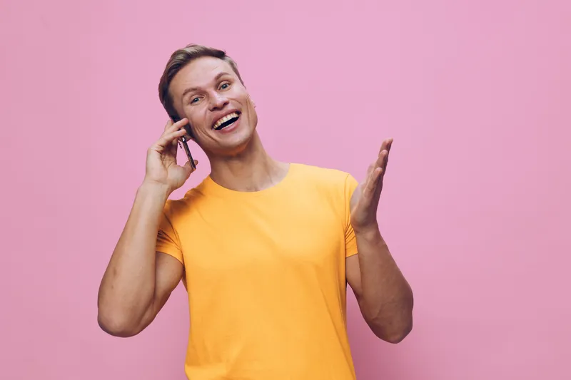 Smiling woman wearing bright yellow t-shirt talking on mobile phone and expressing happiness isolated on pink background. People lifestyle cheerful positive emotions casual summer style.