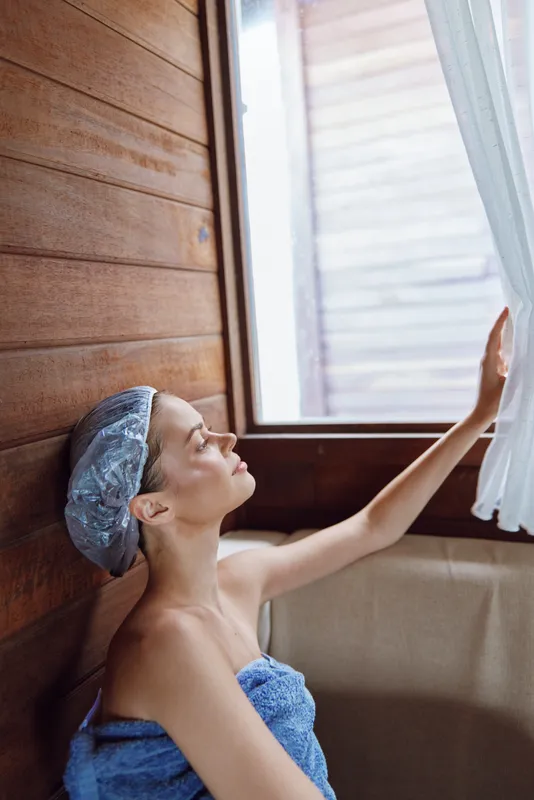Relaxed woman in bathrobe and shower cap enjoys natural light by wooden sauna window, embracing calm and peaceful wellness moment indoors.