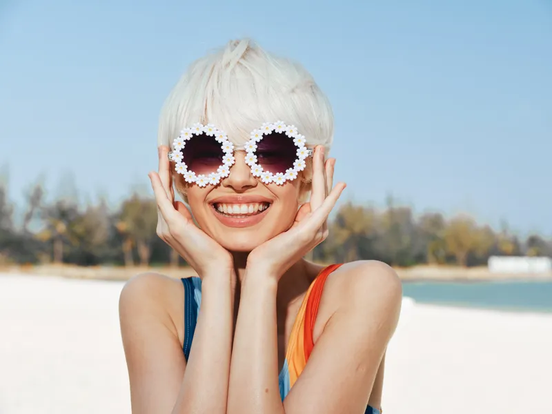Young woman with platinum hair wearing flower sunglasses at a sunny beach, smiling and resting her face on her hands. Bright, playful summer vibe with casual seaside style.