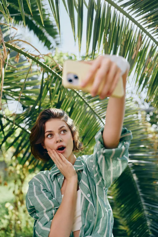 Young woman takes a selfie outdoors among palm leaves, surprised expression, sunny day, casual striped shirt, vibrant green foliage.