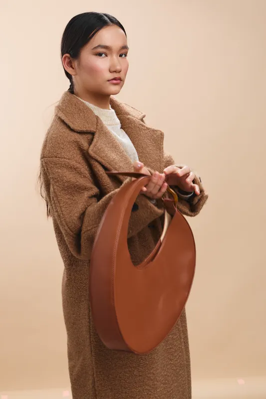 Asian woman in a warm coat holds a circular handbag against a neutral backdrop, presenting a poised and stylish professional image with calm confidence.