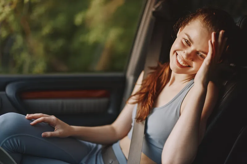 Young woman in casual sportswear smiling joyfully in a car, reflecting happiness and comfort, surrounded by greenery in a vibrant setting