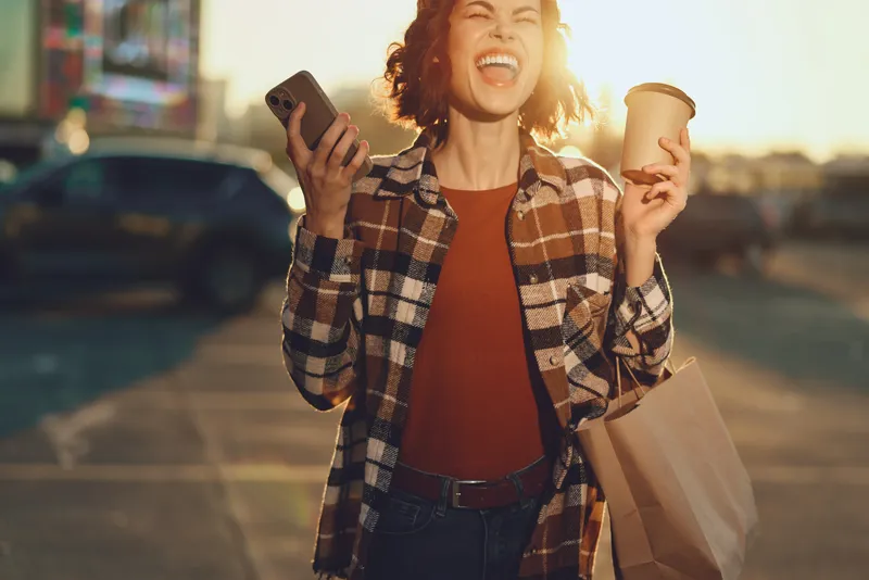 Woman with coffee and phone smiling while shopping in urban sunlight, a candid lifestyle portrait at golden hour glow. Authenticity and mindful living enhance emotional storytelling and joy