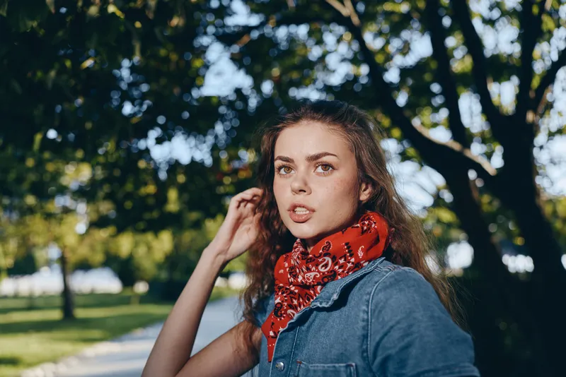 Lifestyle woman in modern boho-western denim street style with a red bandana, natural makeup, and film color tones in an outdoor park setting during daytime.