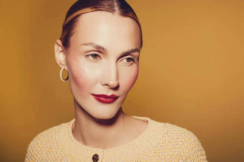 Portrait of a woman with red lipstick and gold earrings wearing a beige sweater against a warm yellow background. Concept of beauty, fashion, and elegance in studio lighting.