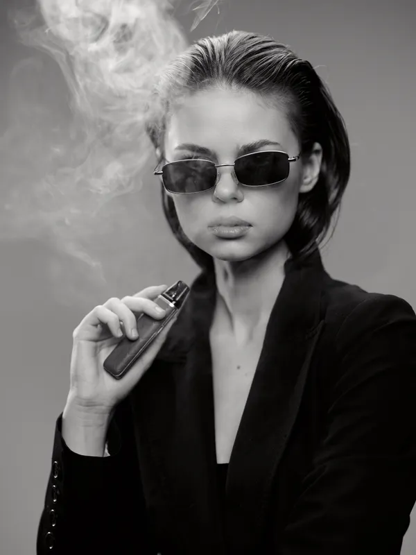 Black and white portrait of a woman vaping in sunglasses, wearing a dark jacket with smoke around her face
