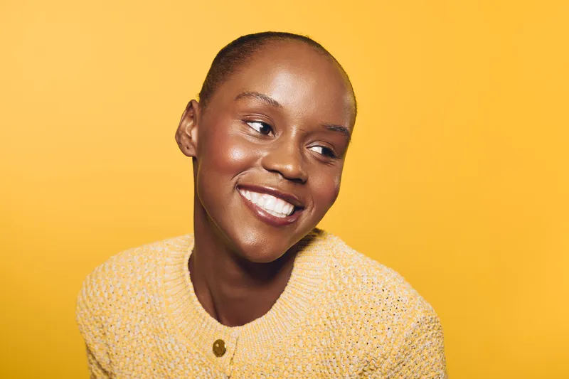 Smiling young black woman with dark skin wearing a yellow sweater looks away on yellow background. Concept of happiness, natural beauty, and positive mood in studio portrait.