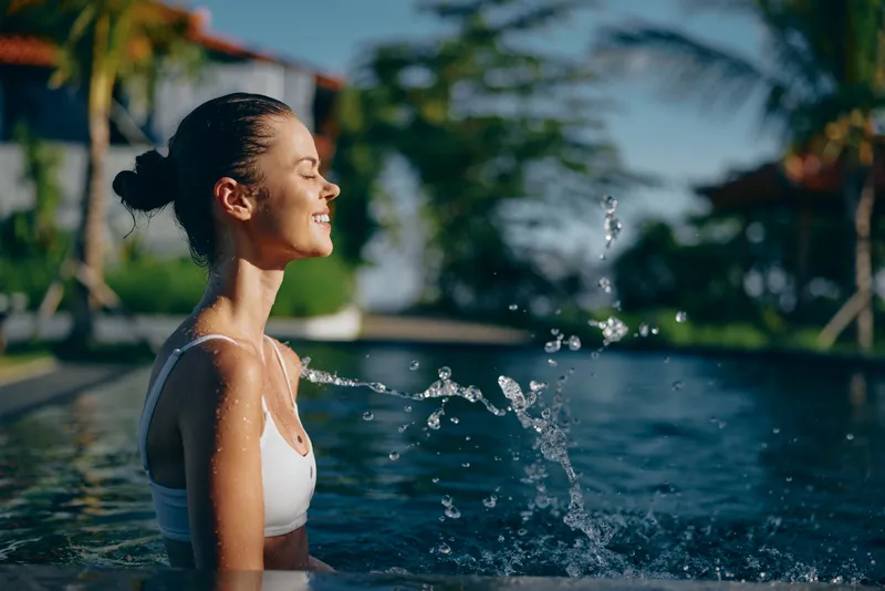 A young woman in a white swimsuit enjoys a sunny day in a swimming pool with water splashing around her. The tropical background creates a relaxing lifestyle atmosphere.