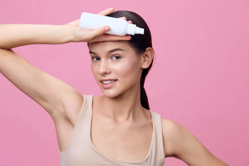 Young woman applying hair product on her forehead, smiling confidently against a pink background, showcasing healthy hair care routine