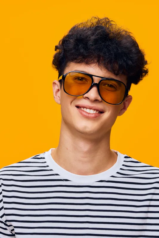 smiling man with curly hair wearing striped shirt and large orange sunglasses against bright yellow background, youthful and cheerful portrait