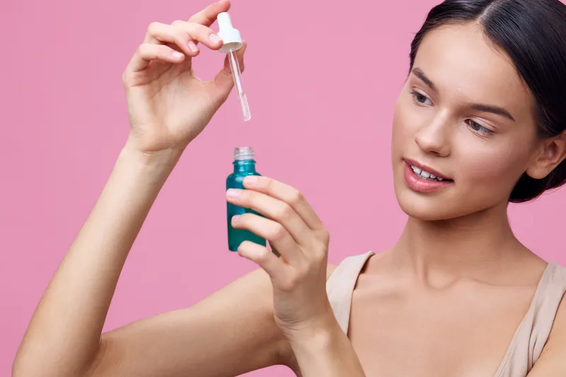 A young woman holding a dropper above a green bottle, showcasing a skincare serum against a pink background, exuding confidence and curiosity in her expression