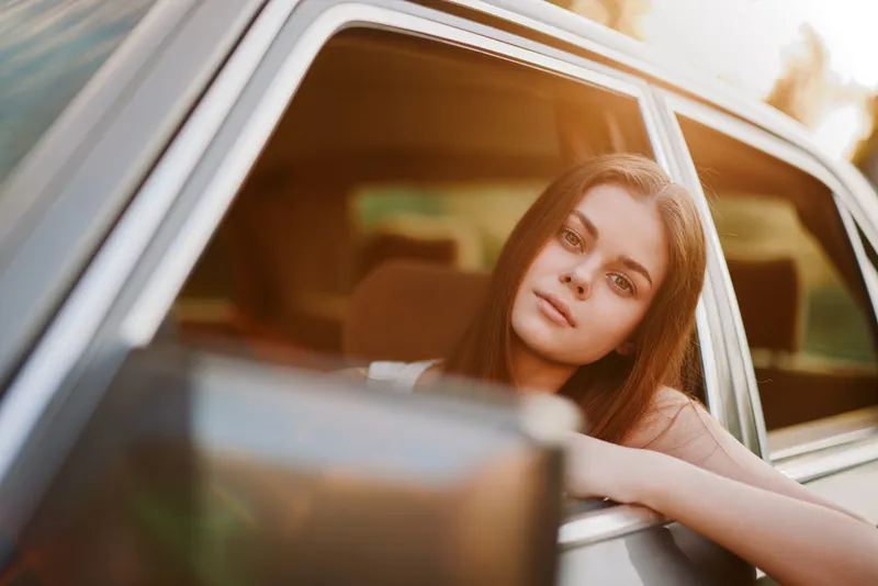Young woman enjoying a sunny day, leaning out of a car window with a relaxed expression, wearing casual summer clothing, highlighting a carefree spirit.