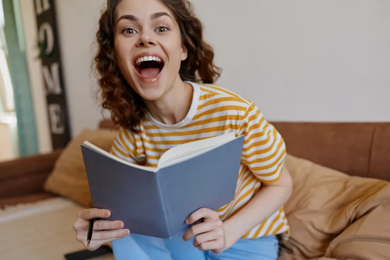 A joyful young woman sits on a couch, reading a blue notebook, her mouth wide open in surprise and a bright smile lighting up the cozy living room.