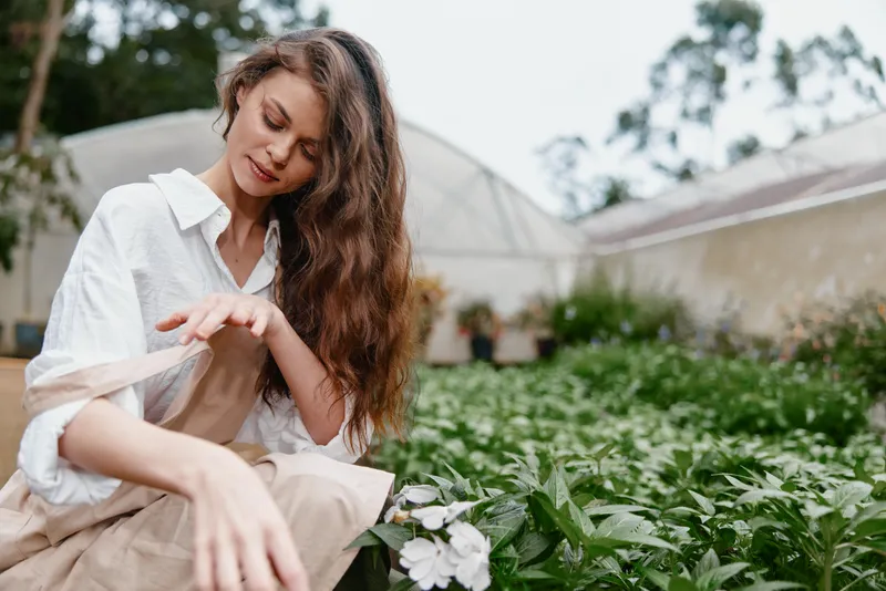 Natural woman in a garden wearing a white shirt and beige apron, showcasing a joyful expression among vibrant plants and flowers during spring