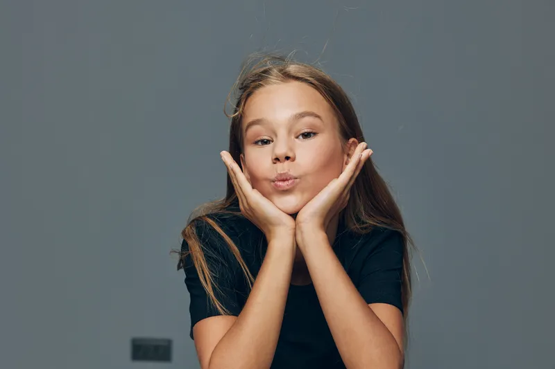 Girl with long hair wearing black shirt posing with hands on face and playful expression on gray background in studio. Child, beauty and casual style with confident look.
