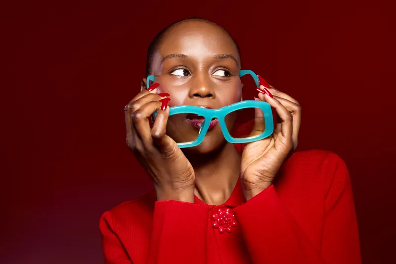 Portrait of a dark skinned African woman holding large turquoise glasses near her face wearing a red sweater on a red background. Concept of fashion, style, and beauty.