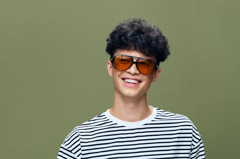Smiling man with curly hair and orange sunglasses wearing a striped shirt, standing against a plain green background, radiating cheerful and casual vibe.