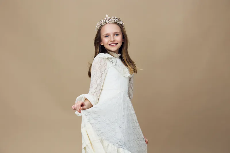 Young girl in a beautiful lace dress with a crown, smiling joyfully, set against a soft beige background, perfect for themes of childhood and celebration