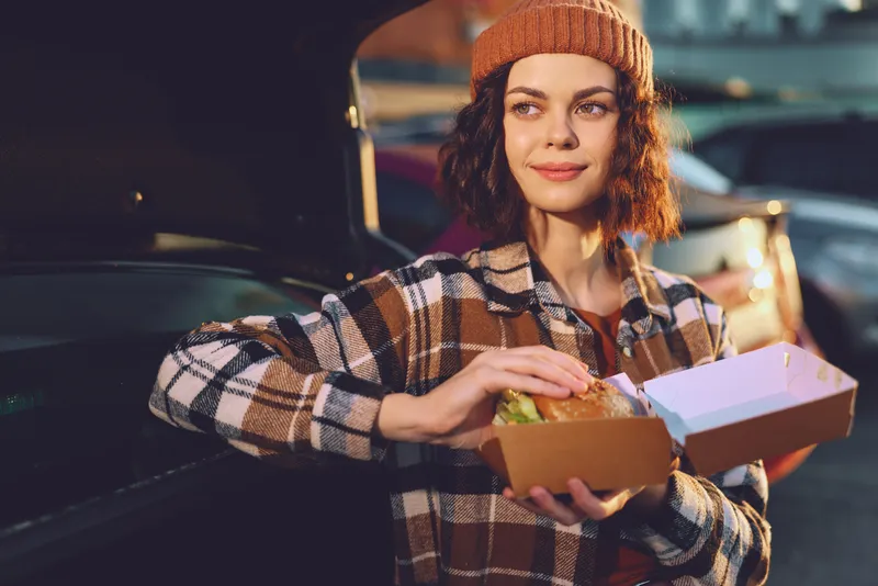 Woman eating burger from car trunk at evening, beanie and plaid jacket, candid smile and authenticity in a golden hour glow, mindful living and emotional storytelling urban scene.