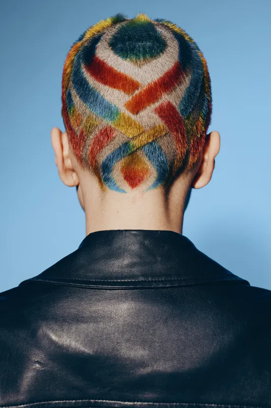Woman with colorful shaved hair design viewed from the back, wearing a black leather jacket against a blue background, showcasing bold artistic hairstyle and fashion.