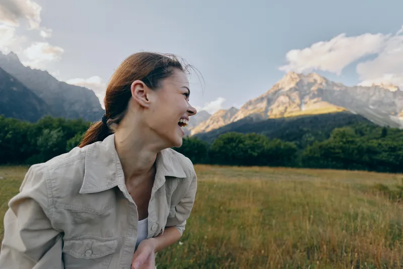 Smiling woman with short hair enjoying nature outdoors in a field with mountains in the background. Concept of lifestyle, happiness, and freedom in natural light.