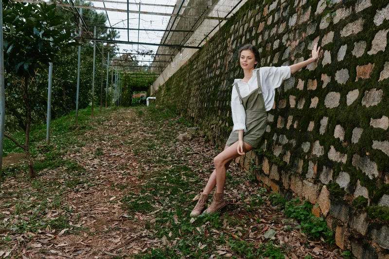 Young woman in olive green dress and white blouse posing against textured stone wall in nature, conveying relaxed and serene emotions, perfect for seasonal lifestyle themes