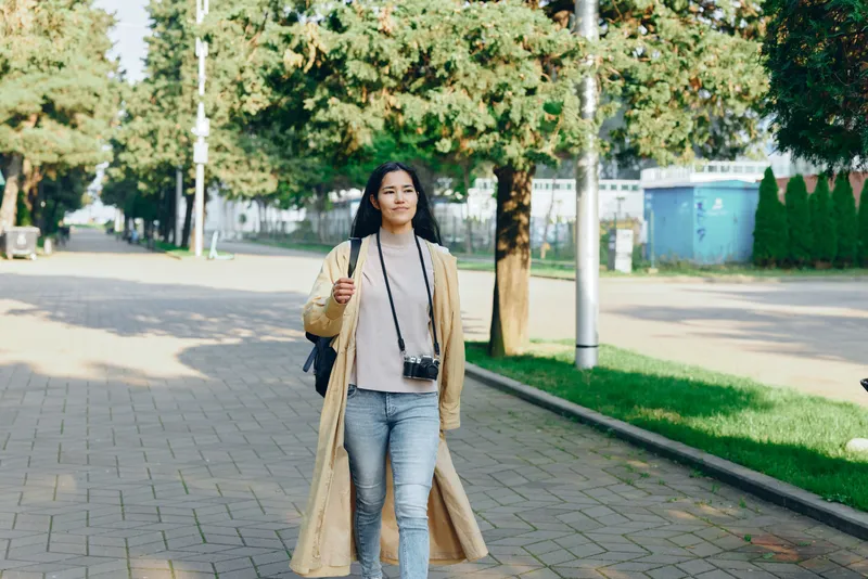 Woman walking on a tree-lined street with a camera around her neck, wearing a long beige coat and jeans, carrying a bag, daytime urban lifestyle scene