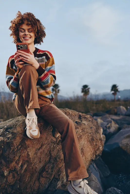 Outdoor lifestyle portrait of woman with curly hair smiling while using smartphone and sitting on large rock, casual striped sweater and jeans, bright sky, relaxed travel mood