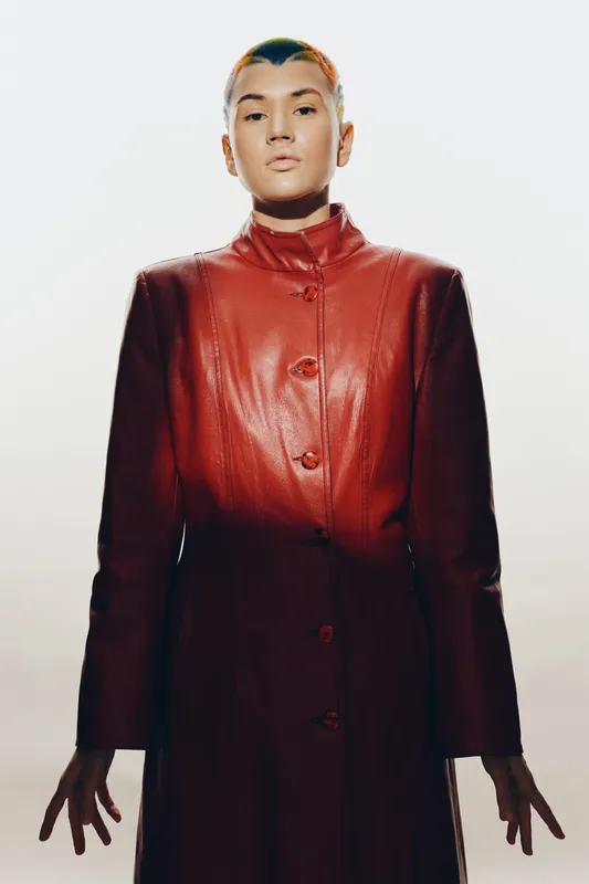 Woman with short hair wearing a red leather coat, standing confidently against a bright background, showcasing bold fashion and modern style.