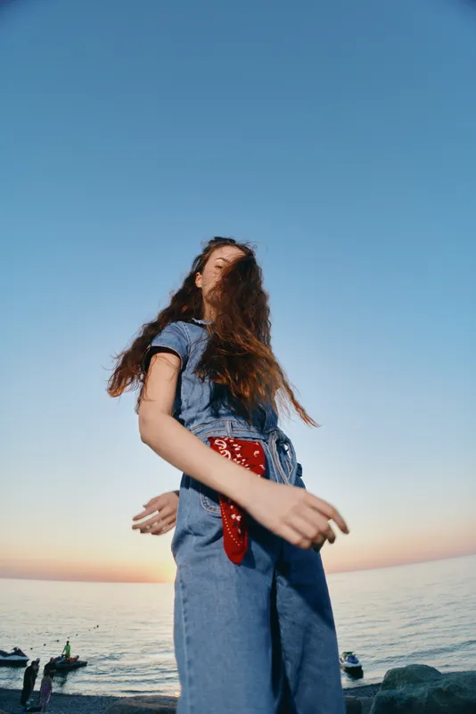 Lifestyle woman in modern boho-western denim street style standing by the sea at sunset with warm film color tones creating a natural and relaxed vibe.