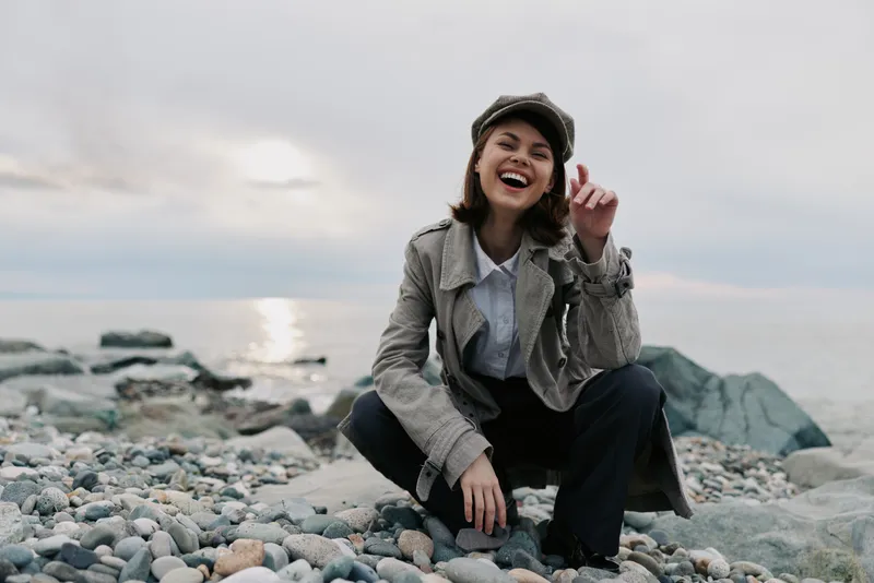 Smiling young woman in stylish coat and hat squats on rocky beach near calm sea under cloudy sky. Concept of happiness, fashion, outdoor lifestyle, and natural beauty by the water.