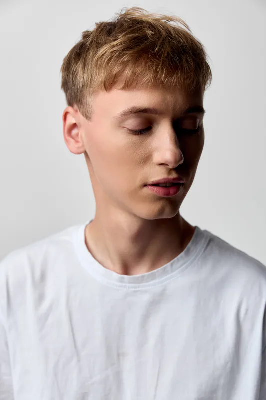 Calm, serene youth with a soft gaze and eyesclosed, wearing a plain white shirt. A minimalist studio setting uses soft lighting to convey contemplation, gentle mood, and quiet confidence in a clean,