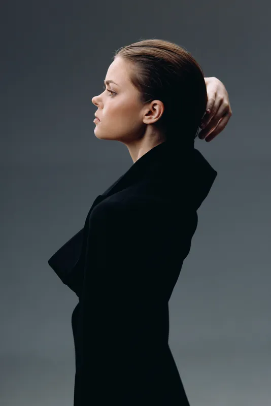 Profile portrait of a serious woman in black dress with neutral background, showing elegance and calm expression.
