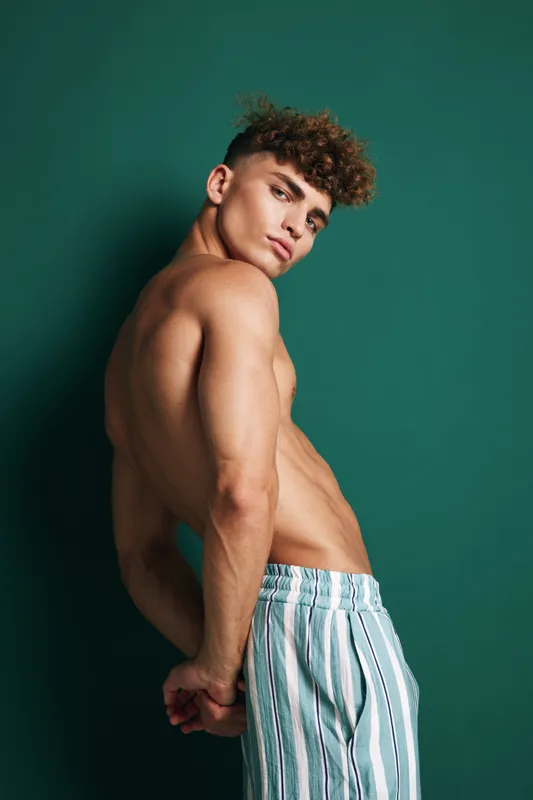 Young muscular man with curly hair posing shirtless against dark green background, showing toned torso and arms with striped casual shorts, looking confidently at camera.