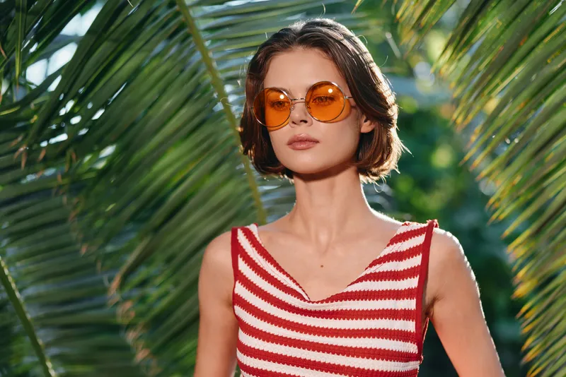 A stylish woman wearing a red and white striped top and orange sunglasses stands among lush palm leaves, projecting a chic tropical vibe and confident mood.
