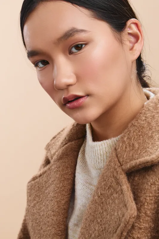 Asian woman with a calm expression wearing a wool coat and knit sweater, close up portrait in a warm studio setting with soft lighting highlighting natural features.