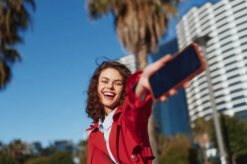 Young woman with brunette hair takes a sunny selfie outdoors in an urban setting, wearing a red jacket and denim, smiling in a bright, energetic moment.