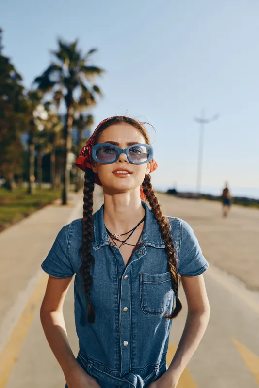 young woman with braids wearing sunglasses and denim dress walking on sunny street near palm trees with blue sky background