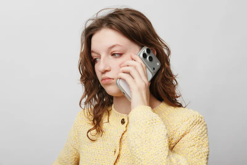 Young woman with curly hair wearing yellow sweater talks on smartphone, looking away with serious expression. Concept of communication, technology, and thoughtful mood.