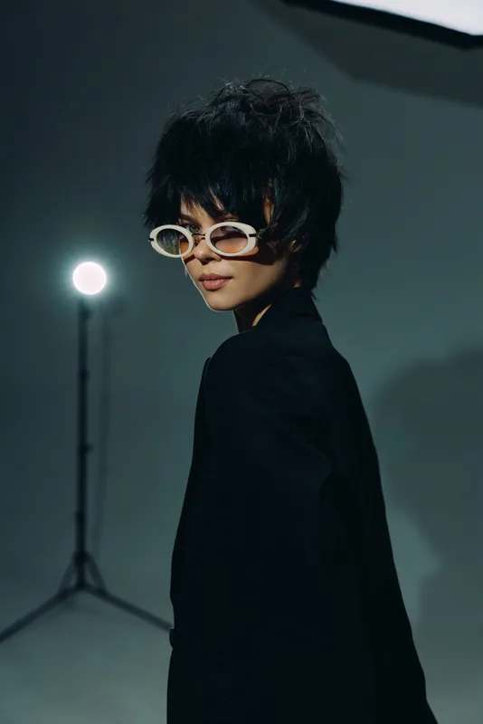 Stylish woman with short dark hair wearing trendy white sunglasses in dark studio setting, modern fashion look