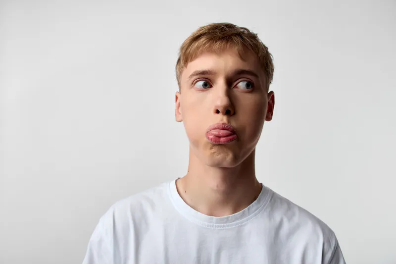Young man with a playful face sticking his tongue out, wearing a white t shirt, in a studio with a neutral background, conveying a casual humorous mood and lighthearted charm, captured in a clean