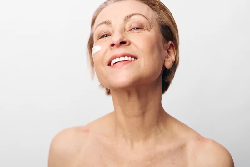 Smiling mature woman with natural skin applies cream on cheek. Close-up on white background. Concept of skincare, aging beauty, self-care, wellness, and healthy lifestyle for women.
