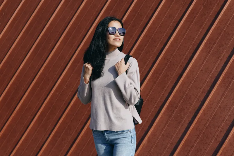 Woman with sunglasses and backpack posing against a diagonal wooden wall in casual chic attire with a soft sweater and jeans, urban street fashion, confident contemporary style.