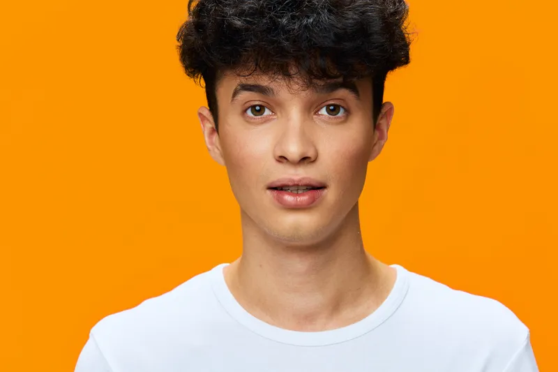 young man with curly hair wearing a white t-shirt standing against bright orange background, looking directly with neutral expression, studio portrait