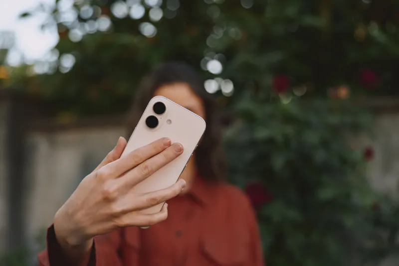 A woman in casual attire holds a smartphone for a selfie outdoors, with a blurred garden background. A lifestyle moment emphasizing connection, self expression, and everyday tech